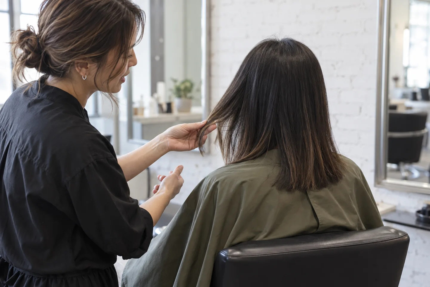 Colorist lifting a dry side section of an adult client’s shoulder-length dark hair in a white-brick salon