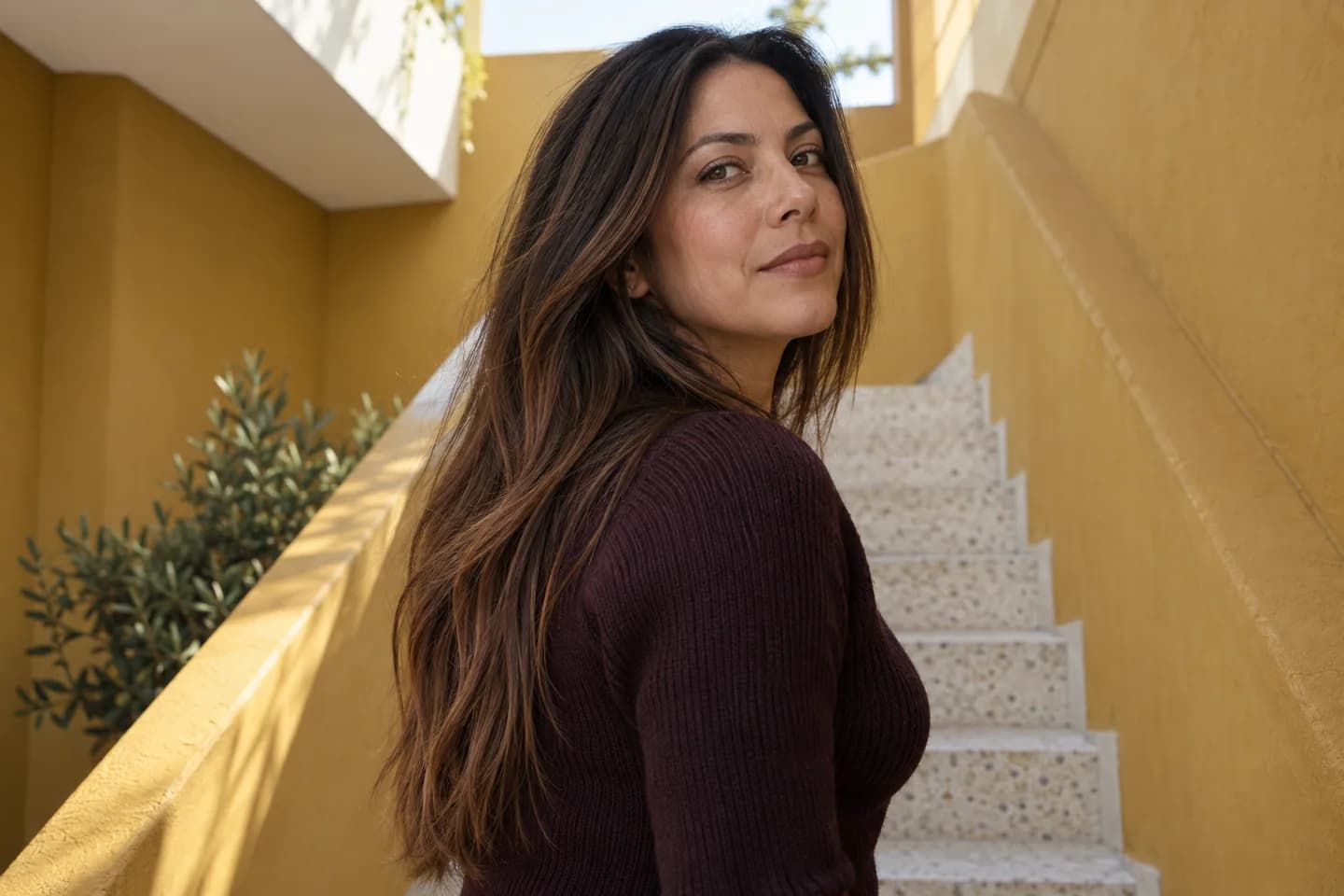 Adult woman with long chestnut hair and restrained copper ribbons on a mustard staircase, wearing a plum knit top