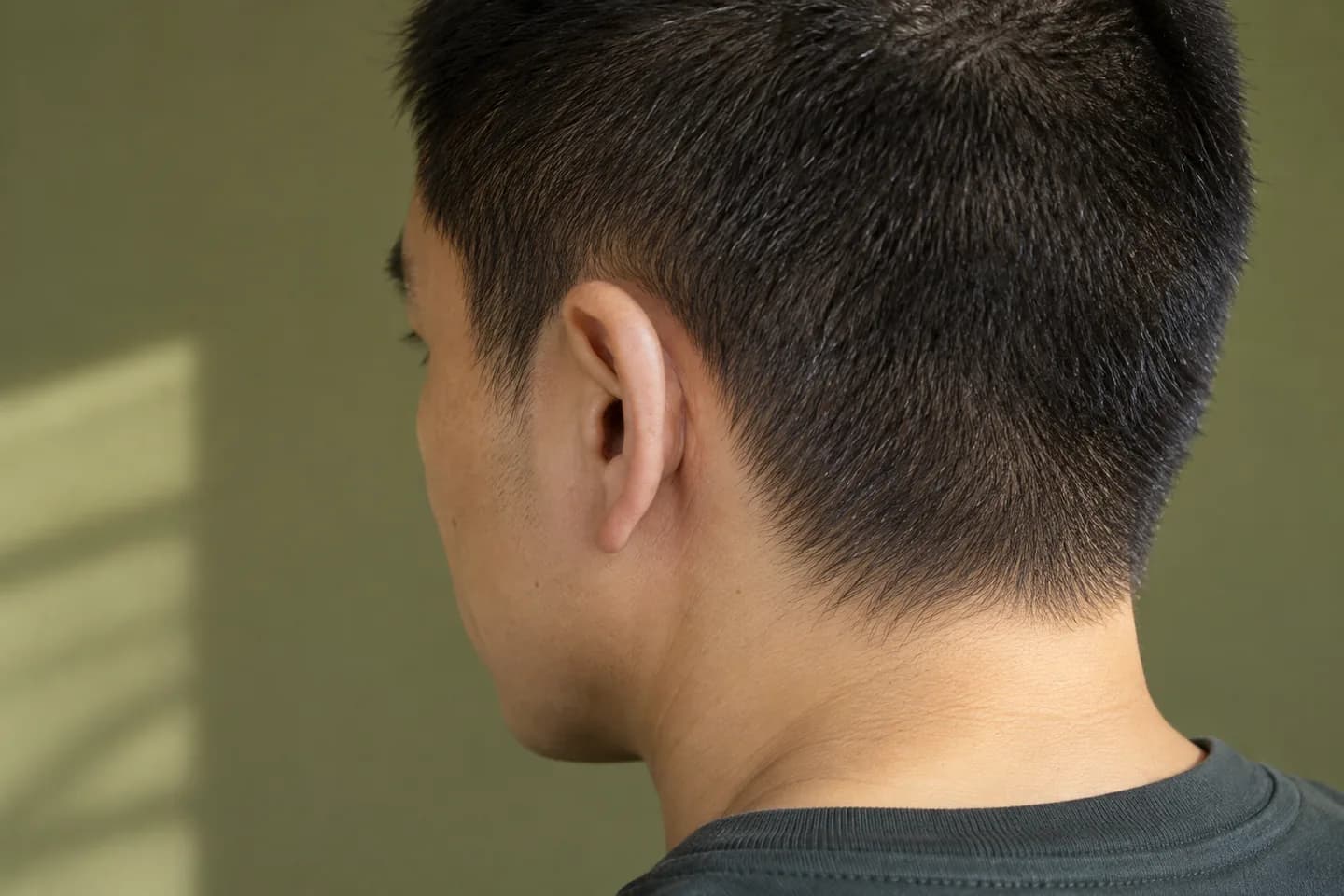Nahansicht eines dunklen Buzz Cuts von schräg hinten mit weicher natürlicher Kontur an Ohr und Nacken vor einer olivfarbenen Wand