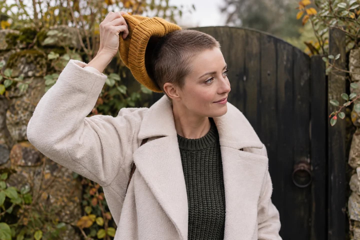 Erwachsene Frau mit dunkelblondem Buzz Cut nimmt an einem hölzernen Gartentor eine senfgelbe Strickmütze ab