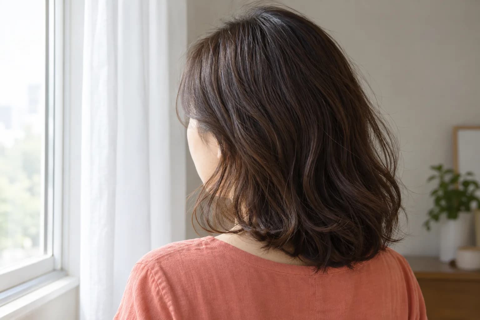 Rückenansicht einer erwachsenen Frau mit dunkelbraunen Wellen neben einem weißen Vorhang im indirekten Tageslicht