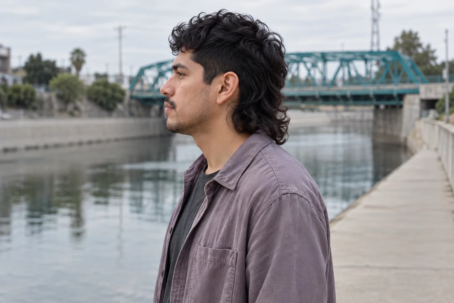 Homem adulto com mullet preto ondulado e sobrecamisa lilás de perfil ao lado de uma ponte verde-azulada sobre um canal