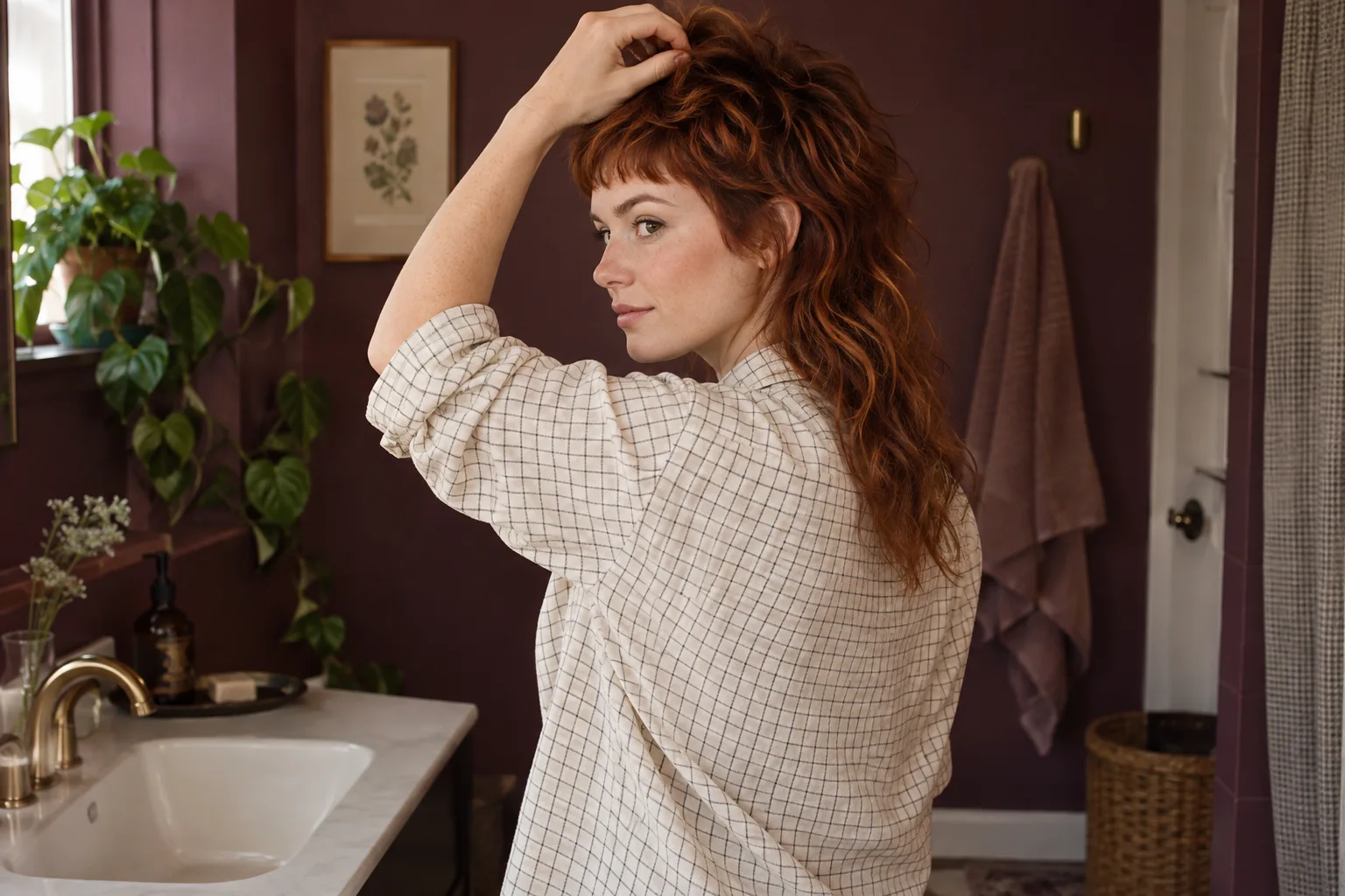 Mulher adulta com mullet acobreado ondulado ajeita levemente o topo seco em um banheiro cor de ameixa
