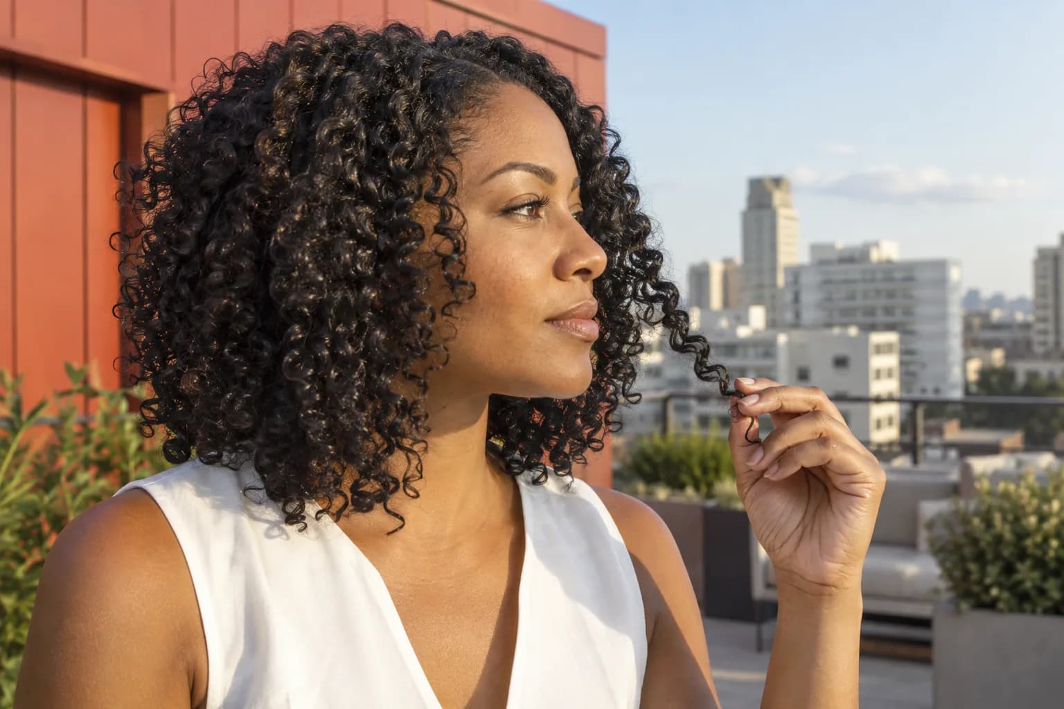 Adult woman with dark shoulder-length spiral curls gently holding one curl end on a rooftop