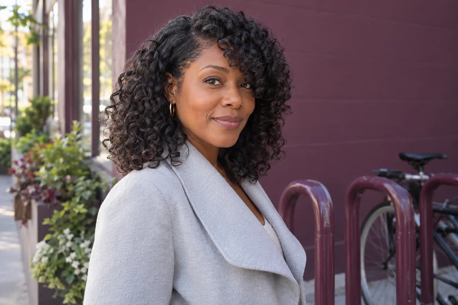 Adult Black woman with loose dark curls and an asymmetrical curly fringe beside a plum bicycle rack
