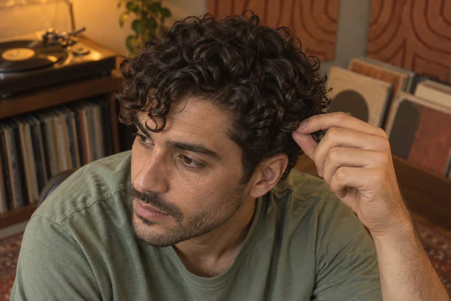 Adult man in a record room gently lifting a dark curl above his temple to show the connection between his top and sides